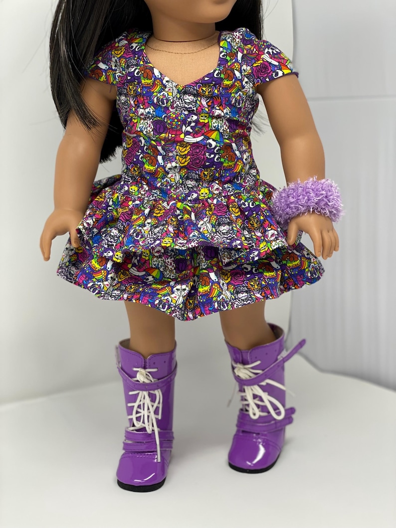 K&ouml;nnte beinhalten: Eine Puppe in einem farbenfrohen Kleid mit Totenkopf- und Regenbogenmuster, einem flauschigen lila Armband und passenden lila Stiefeln mit wei&szlig;en Schn&uuml;rsenkeln. Das Kleid hat einen ger&uuml;schten Rock und kurze &Auml;rmel. Die Stiefel haben ein gl&auml;nzendes Finish.