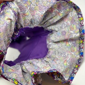 K&ouml;nnte beinhalten: Ein farbenfrohes Stoffteil mit lila Innenfutter. Der Au&szlig;enstoff weist ein lebhaftes Muster aus Totenk&ouml;pfen, Rosen und anderen skurrilen Designs in verschiedenen Farben auf. Der Artikel wird offen gehalten und gibt den Blick auf das Innere frei.
