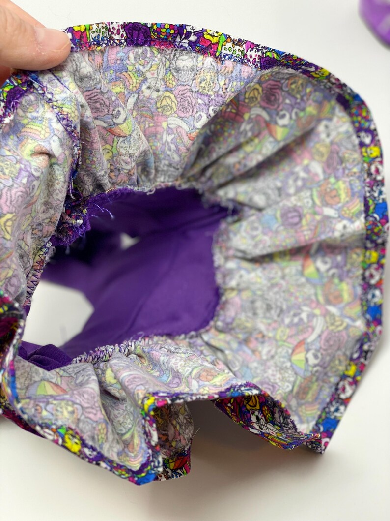 K&ouml;nnte beinhalten: Ein farbenfrohes Stoffteil mit lila Innenfutter. Der Au&szlig;enstoff weist ein lebhaftes Muster aus Totenk&ouml;pfen, Rosen und anderen skurrilen Designs in verschiedenen Farben auf. Der Artikel wird offen gehalten und gibt den Blick auf das Innere frei.