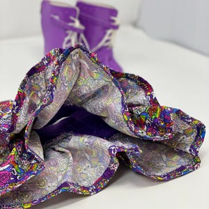 K&ouml;nnte beinhalten: Ein bunter Rock mit Muster und lila Futter und Besatz. Im Hintergrund sind zwei lila Gummistiefel mit wei&szlig;en Schn&uuml;rsenkeln zu sehen, was auf ein verspieltes, jugendliches Thema hindeutet.