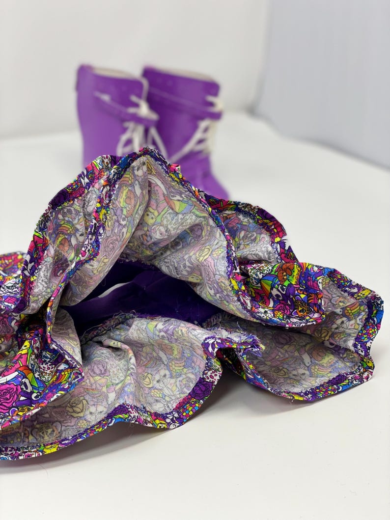 K&ouml;nnte beinhalten: Ein bunter Rock mit Muster und lila Futter und Besatz. Im Hintergrund sind zwei lila Gummistiefel mit wei&szlig;en Schn&uuml;rsenkeln zu sehen, was auf ein verspieltes, jugendliches Thema hindeutet.