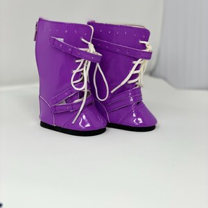 K&ouml;nnte beinhalten: Ein Paar gl&auml;nzende, lila Puppenstiefel mit wei&szlig;en Schn&uuml;rsenkeln und Schnallen. Die Stiefel haben eine gl&auml;nzende Oberfl&auml;che und schwarze Sohlen. Das Design umfasst Riemen und einen Rei&szlig;verschluss auf der R&uuml;ckseite. Diese Stiefel sind f&uuml;r Puppen.