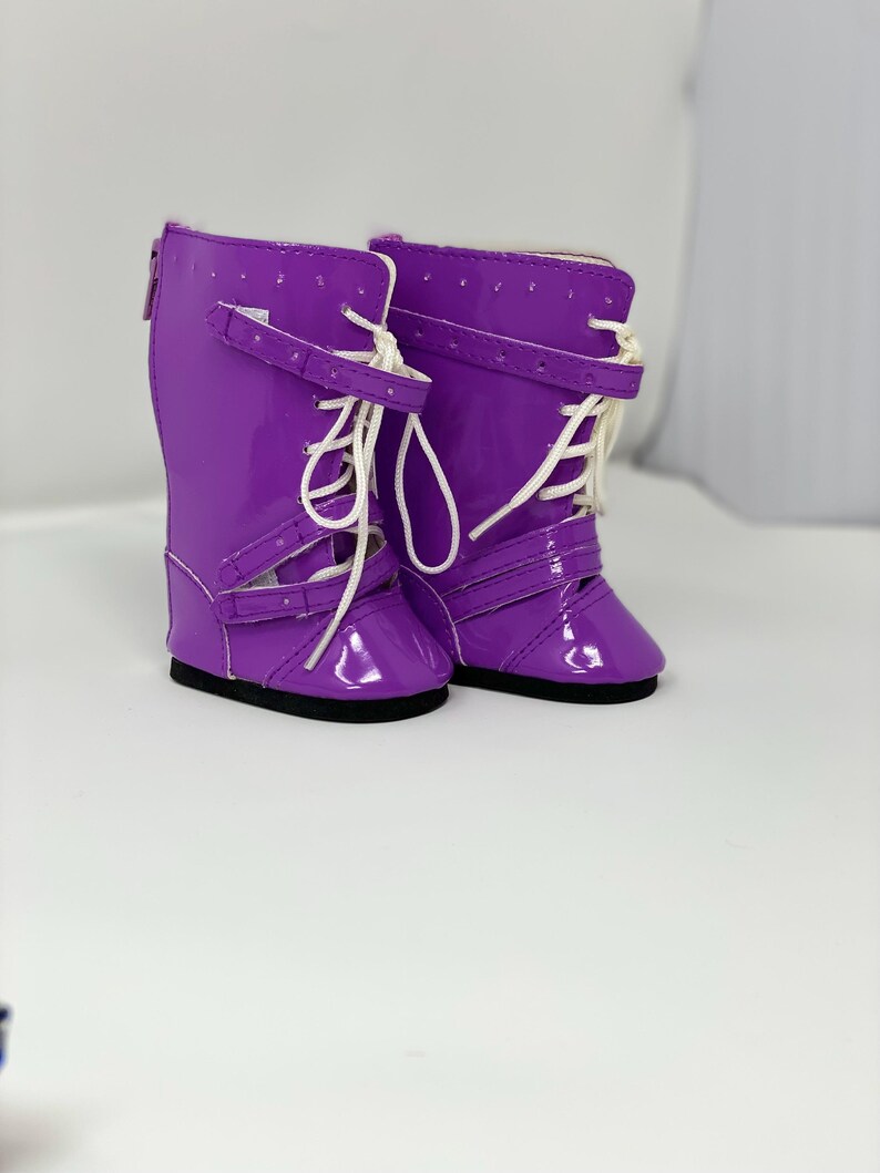 K&ouml;nnte beinhalten: Ein Paar gl&auml;nzende, lila Puppenstiefel mit wei&szlig;en Schn&uuml;rsenkeln und Schnallen. Die Stiefel haben eine gl&auml;nzende Oberfl&auml;che und schwarze Sohlen. Das Design umfasst Riemen und einen Rei&szlig;verschluss auf der R&uuml;ckseite. Diese Stiefel sind f&uuml;r Puppen.