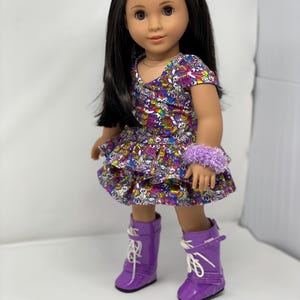 K&ouml;nnte beinhalten: Eine Puppe mit langen schwarzen Haaren, die ein bunt gemustertes Kleid, ein lila Armband und passende lila Stiefel tr&auml;gt. Das Kleid hat einen ger&uuml;schten Rock. Rosa Haarspangen in Blumenform schm&uuml;cken das Haar der Puppe.