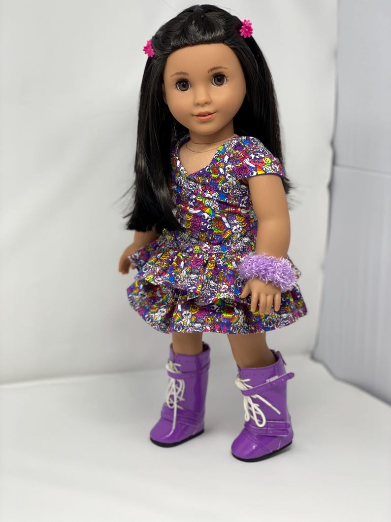 K&ouml;nnte beinhalten: Eine Puppe mit langen schwarzen Haaren, die ein bunt gemustertes Kleid, ein lila Armband und passende lila Stiefel tr&auml;gt. Das Kleid hat einen ger&uuml;schten Rock. Rosa Haarspangen in Blumenform schm&uuml;cken das Haar der Puppe.