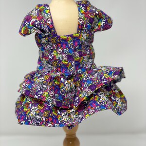 K&ouml;nnte beinhalten: Ein mehrfarbiges Hundekleid mit quadratischem Ausschnitt und R&uuml;schenrock. Das Kleid ist mit einem sich wiederholenden Muster aus Cartoon-Charakteren in Lila, Blau, Rosa und Gelb bedeckt. Pr&auml;sentiert auf einer Holzpuppe.