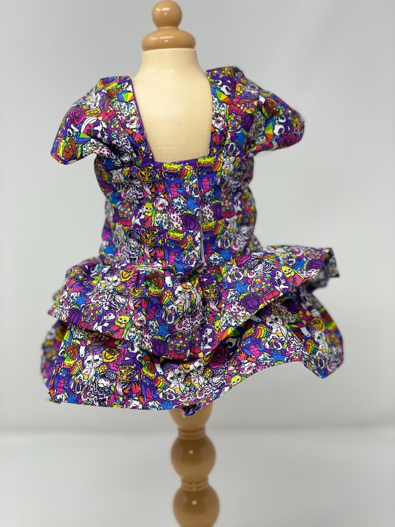 K&ouml;nnte beinhalten: Ein mehrfarbiges Hundekleid mit quadratischem Ausschnitt und R&uuml;schenrock. Das Kleid ist mit einem sich wiederholenden Muster aus Cartoon-Charakteren in Lila, Blau, Rosa und Gelb bedeckt. Pr&auml;sentiert auf einer Holzpuppe.