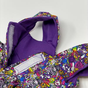 K&ouml;nnte beinhalten: Nahaufnahme eines Haustierzubeh&ouml;rs mit einem lebendigen, farbenfrohen Muster. Das Zubeh&ouml;r hat eine lila Innenseite und eine gemusterte Au&szlig;enseite mit Totenk&ouml;pfen, Blumen und anderen Designs. Wei&szlig;e Klettverschl&uuml;sse sind sichtbar.