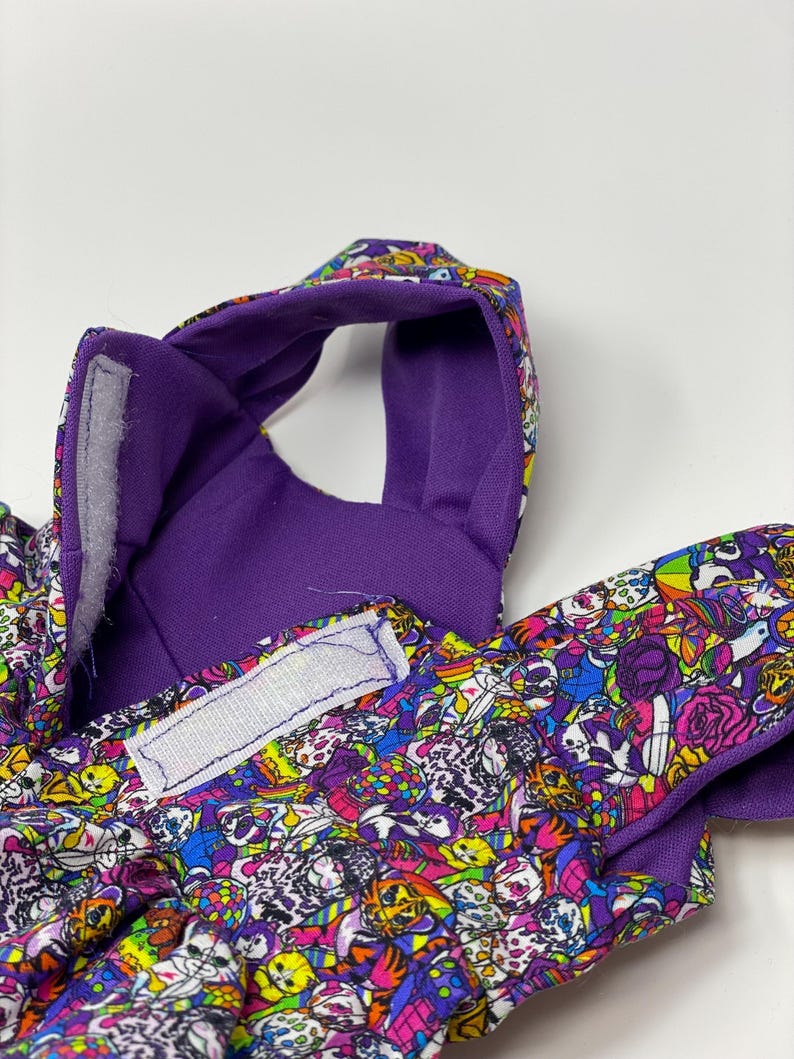 K&ouml;nnte beinhalten: Nahaufnahme eines Haustierzubeh&ouml;rs mit einem lebendigen, farbenfrohen Muster. Das Zubeh&ouml;r hat eine lila Innenseite und eine gemusterte Au&szlig;enseite mit Totenk&ouml;pfen, Blumen und anderen Designs. Wei&szlig;e Klettverschl&uuml;sse sind sichtbar.