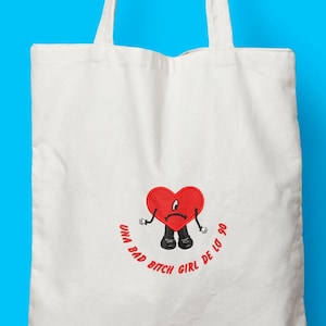 May include: White canvas tote bag featuring a red heart-shaped character with a sad expression. The character has black shoes and arms. The text "UNA BAD BITCH GIRL DE LO 90" is printed in red around the character. The bag has two white handles.