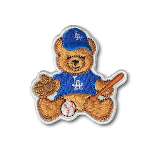 May include: Embroidered patch featuring a brown teddy bear wearing a blue baseball cap and jersey with "LA" lettering. The bear holds a baseball bat and mitt, with a baseball at its feet. The patch has a white border.