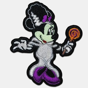 May include: A black and white embroidered patch of a cartoon character dressed as a monster holding a lollipop. The character has green skin, purple shoes, and a purple bow. The lollipop is orange and purple.