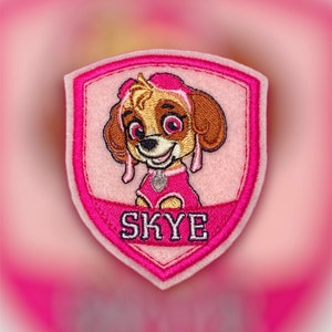 May include: Pink and white embroidered patch featuring a cartoon dog with brown fur, pink ears, and a pink collar. The dog is wearing a pink and white outfit and has a pink bow on its head. The patch has the word "SKYE" embroidered in white letters on a pink background.