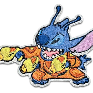 May include: A blue alien character with large ears and a toothy grin, wearing an orange jumpsuit and holding two yellow objects in its hands. The character is embroidered on a white background.