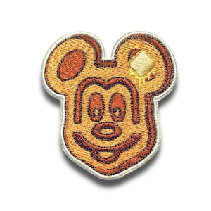 May include: Embroidered patch in the shape of a Mickey Mouse pancake with a pat of butter. The patch is tan and brown with a white border. The butter is yellow with a dripping effect. The patch is on a white background.