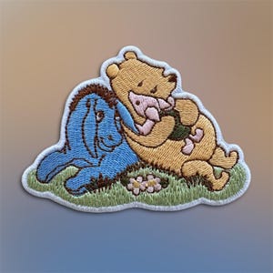 May include: Embroidered patch featuring Winnie the Pooh, Piglet, and Eeyore. Pooh hugs Piglet, while Eeyore lies nearby. The patch has a white border, with green grass and small flowers at the bottom. The characters are in their classic colors.