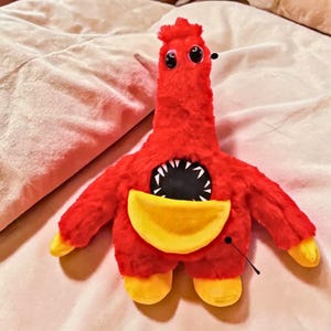 May include: A plush toy in the shape of a red creature with a long neck, large black eyes, and a yellow pouch. The creature has a wide, toothy mouth and yellow feet and hands. The toy is made of soft, fuzzy material.