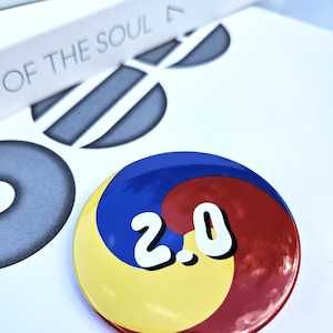 May include: A round button with a yellow, red, and blue design, featuring the number "2.0" in white. The button is set against a white background with a book and graphic elements.
