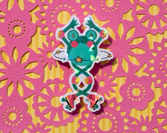 May include: A holographic sticker of a green frog wearing roller skates and orbiting a planet. The frog has a pink nose and yellow stars on its body. The planet is blue and green with a yellow star on it.