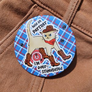 May include: A round button with a cartoon cat wearing a cowboy hat, red bandana, and cowboy boots. The cat is standing on a pink target. Text on the button reads "hold on Partner" and "I'm over-STIMULATED". The background is blue with a plaid pattern.