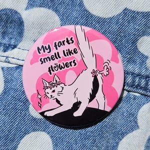 May include: A pink button with a cartoon cat illustration. The cat is white with a flower crown and is depicted with a flower-shaped fart. The text on the button reads "My farts smell like flowers."