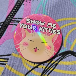May include: A pink button with the text "SHOW ME YOUR KITTIES" and a cartoon cat face. The button has a holographic effect and is set against a purple background with a zipper and colorful design.
