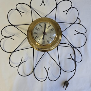 May include: A vintage clock with a gold face and black Roman numeral hour markers. The clock is mounted on a black metal frame with a design of eight hearts. The clock has a black electrical cord with a plug.