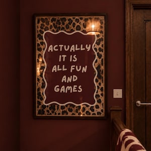 Puede incluir: Una pieza de arte mural rectangular enmarcada con un panel central de color granate oscuro, un borde blanco ondulado y el texto blanco "ACTUALLY IT IS ALL FUN AND GAMES". El fondo detrás del panel de texto muestra un patrón de leopardo marrón y negro.