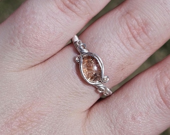 May include: A silver wire wrapped ring with a clear, oval-shaped gemstone with brown and gold inclusions.