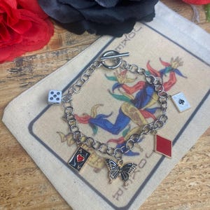 May include: Silver-toned charm bracelet featuring a butterfly, playing card suits, and a die. The bracelet is displayed on a canvas pouch with a joker card design. The bracelet has a toggle clasp.