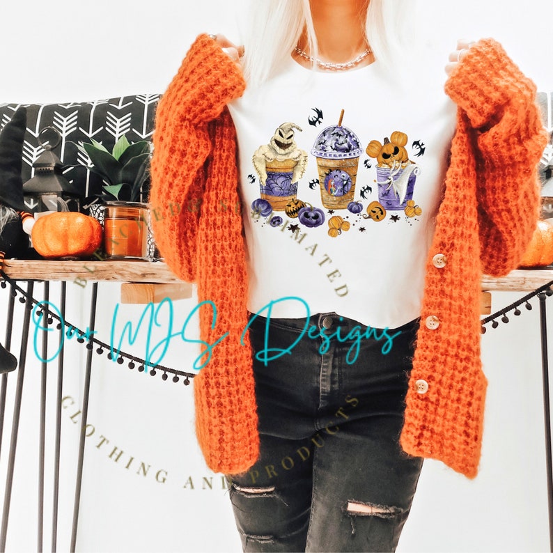 K&ouml;nnte beinhalten: Ein wei&szlig;es T-Shirt mit einem Halloween-Motiv, das drei Comicfiguren in Kaffeetassen zeigt. Die Figuren sind Jack Skellington, Mickey Mouse und ein Geist. Das Design enth&auml;lt Flederm&auml;use, K&uuml;rbisse und das Wort "Animiert".