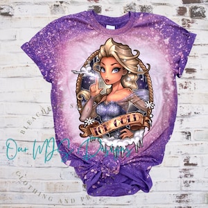 Pode incluir: Camiseta roxa com efeito tie-dye e desbotado, apresentando uma ilustra&ccedil;&atilde;o de desenho animado de uma mulher loira com uma coroa, vestindo um vestido azul e segurando um floco de neve. O texto "ICE COLD" est&aacute; escrito em uma faixa.