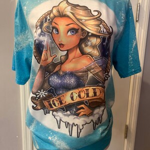 Pode incluir: Uma camiseta azul tie-dye com uma imagem de desenho animado de uma mulher com cabelo loiro e olhos azuis. A mulher est&aacute; vestindo um vestido azul e tem flocos de neve no cabelo. O texto "ICE COLD" est&aacute; escrito em uma faixa abaixo da mulher.