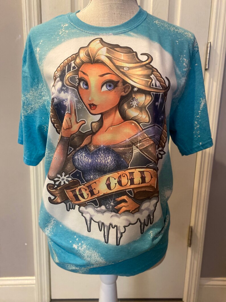 Pode incluir: Uma camiseta azul tie-dye com uma imagem de desenho animado de uma mulher com cabelo loiro e olhos azuis. A mulher est&aacute; vestindo um vestido azul e tem flocos de neve no cabelo. O texto "ICE COLD" est&aacute; escrito em uma faixa abaixo da mulher.