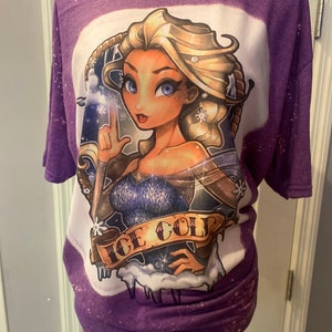 Pode incluir: Camiseta roxa com um design desbotado branco que apresenta uma imagem de desenho animado de uma mulher com cabelo loiro e olhos azuis. A mulher est&aacute; segurando um floco de neve e o texto "ICE COLD" est&aacute; abaixo dela.