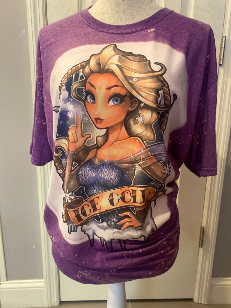 Pode incluir: Camiseta roxa com um design desbotado branco que apresenta uma imagem de desenho animado de uma mulher com cabelo loiro e olhos azuis. A mulher est&aacute; segurando um floco de neve e o texto "ICE COLD" est&aacute; abaixo dela.