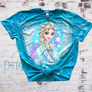 Pode incluir: Uma camiseta azul turquesa com um design salpicado de água sanitária que apresenta uma imagem de desenho animado de Elsa de Frozen. A imagem é cercada por flocos de neve brancos e um fundo azul claro e roxo.