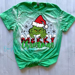 May include: Green t-shirt with a bleached effect featuring a Grinch wearing a Santa hat and the words "Merry Christmas" in red and black buffalo plaid.