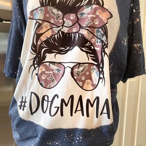 May include: A white t-shirt with a blue and white splatter pattern. The shirt features a graphic of a woman with a messy bun and sunglasses. The bun is decorated with a bow featuring a dog print. The text "#DOGMAMA" is printed below the graphic.