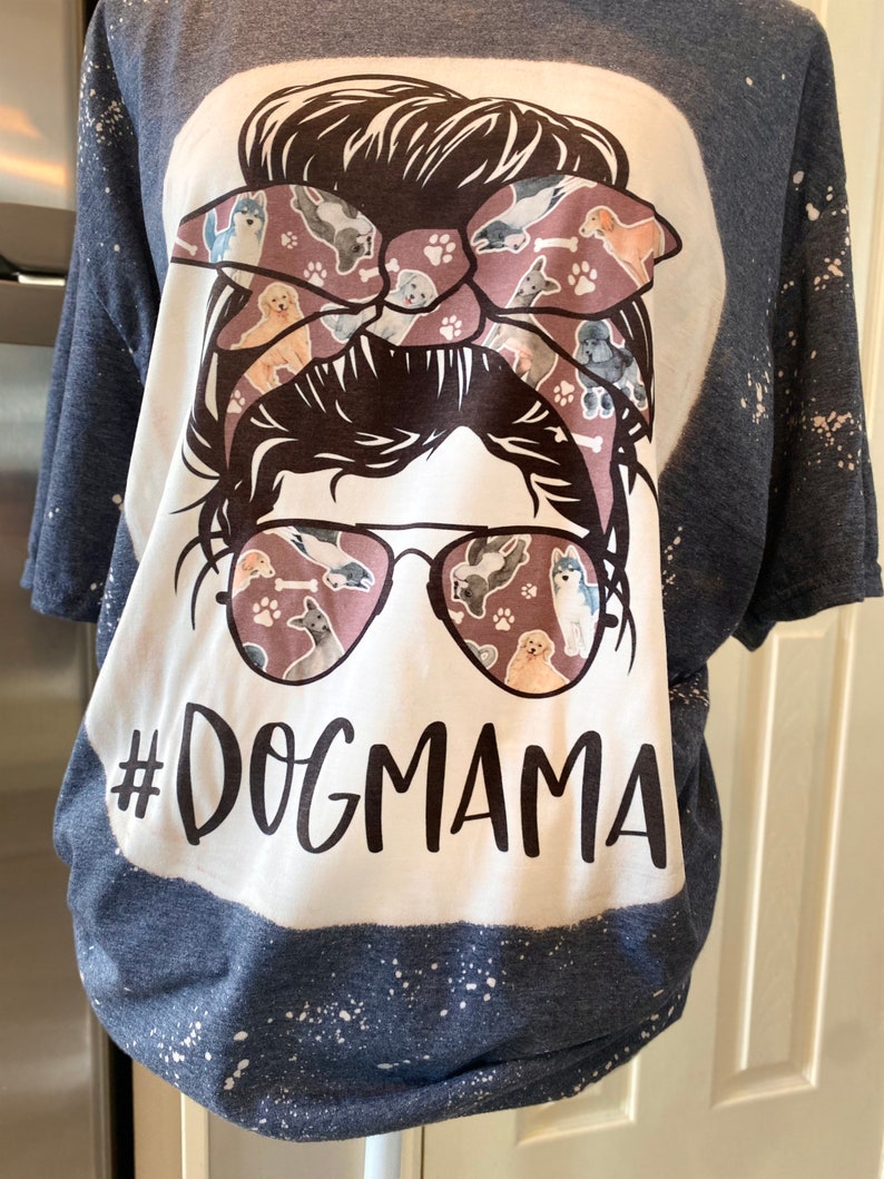 May include: A white t-shirt with a blue and white splatter pattern. The shirt features a graphic of a woman with a messy bun and sunglasses. The bun is decorated with a bow featuring a dog print. The text "#DOGMAMA" is printed below the graphic.