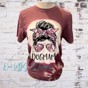 May include: A maroon t-shirt with a bleached design featuring a woman with a messy bun and sunglasses. The sunglasses and headband are decorated with a pattern of dogs. The text "#DogMama" is printed on the shirt.