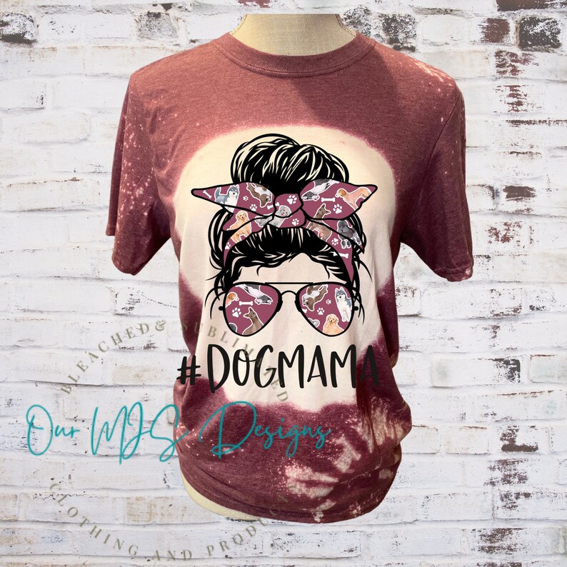 May include: A maroon t-shirt with a bleached design featuring a woman with a messy bun and sunglasses. The sunglasses and headband are decorated with a pattern of dogs. The text "#DogMama" is printed on the shirt.