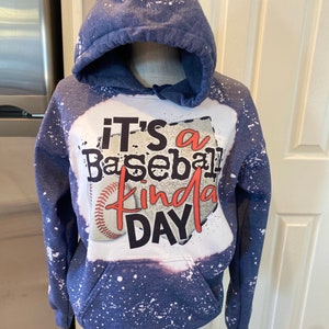 Puede incluir: Una sudadera con capucha azul con salpicaduras de pintura blanca. La parte delantera de la sudadera tiene un estampado gr&aacute;fico de una pelota de b&eacute;isbol y el texto "It's a Baseball Kinda Day".