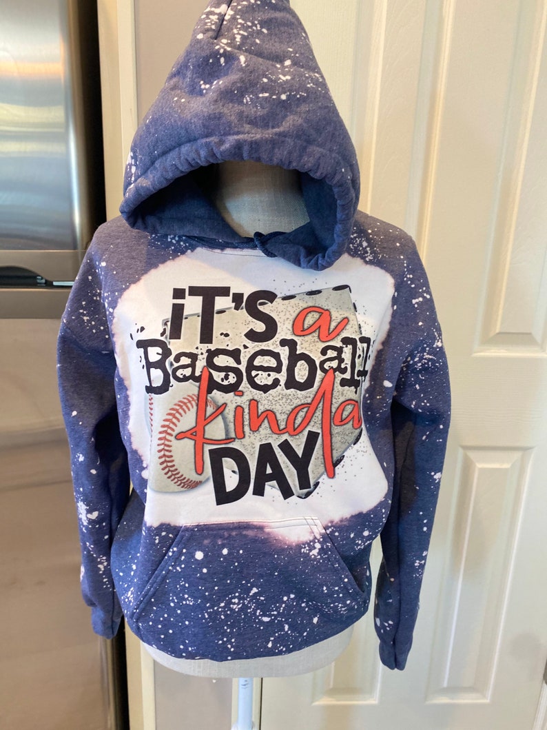 Puede incluir: Una sudadera con capucha azul con salpicaduras de pintura blanca. La parte delantera de la sudadera tiene un estampado gr&aacute;fico de una pelota de b&eacute;isbol y el texto "It's a Baseball Kinda Day".