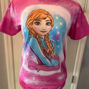 May include: Pink t-shirt with a bleach-splattered design featuring a cartoon image of a blonde girl with blue eyes. The girl is wearing a blue and purple dress and has a pink ribbon around her neck. The background is a light blue with white stars.