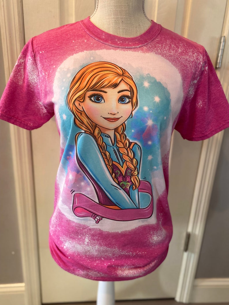 May include: Pink t-shirt with a bleach-splattered design featuring a cartoon image of a blonde girl with blue eyes. The girl is wearing a blue and purple dress and has a pink ribbon around her neck. The background is a light blue with white stars.