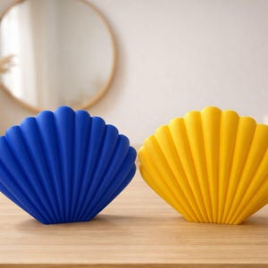 May include: Two seashell-shaped decorative objects, one royal blue and one yellow, sit on a wooden surface. The objects have a ribbed texture and are positioned side-by-side. A round mirror and a vase with dried plants are in the background.
