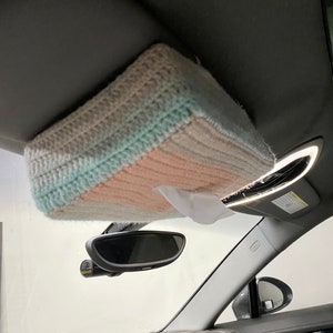 May include: A crocheted tissue box cover in shades of beige, light blue, and pink, hanging from the sun visor of a car.