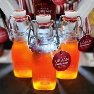 May include: Three glass bottles of honey with a red and white label that says "The Urban Farmhouse Co." The bottles are on a brown surface.