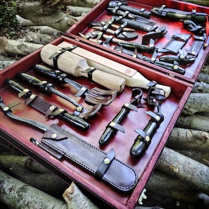 Peut inclure: Une boîte en bois doublée de cuir rouge contenant une sélection d'outils et d'instruments, dont un maillet, des couteaux et une petite hache. Les outils sont disposés en rangées et fixés par des lanières de cuir. La boîte est ouverte et les outils sont visibles.